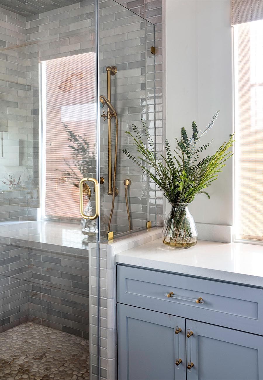 A floor to ceiling shower with gold handles beside white counter tops resting on light blue cabinets